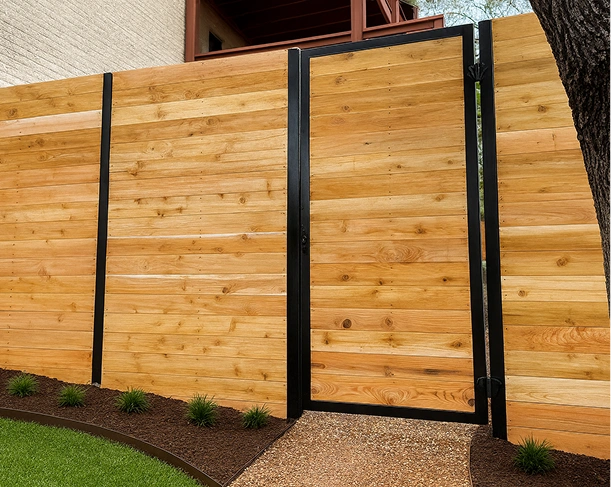A modern horizontal wood privacy fence with warm-toned cedar planks and black metal posts, featuring a matching framed gate that opens onto a pea gravel path.