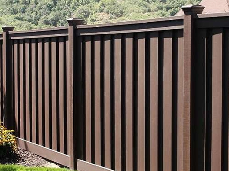 A dark brown vinyl board-on-board privacy fence with decorative post caps bordering a green lawn and mulch bed.
