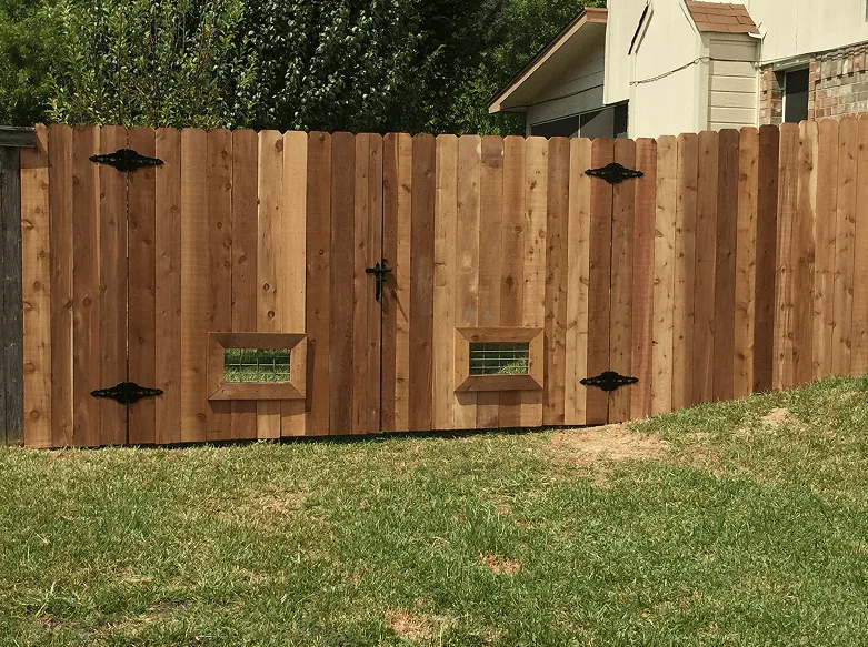 A dog-ear cedar privacy fence with a double gate featuring decorative black metal hinges and small built-in mesh viewing windows.